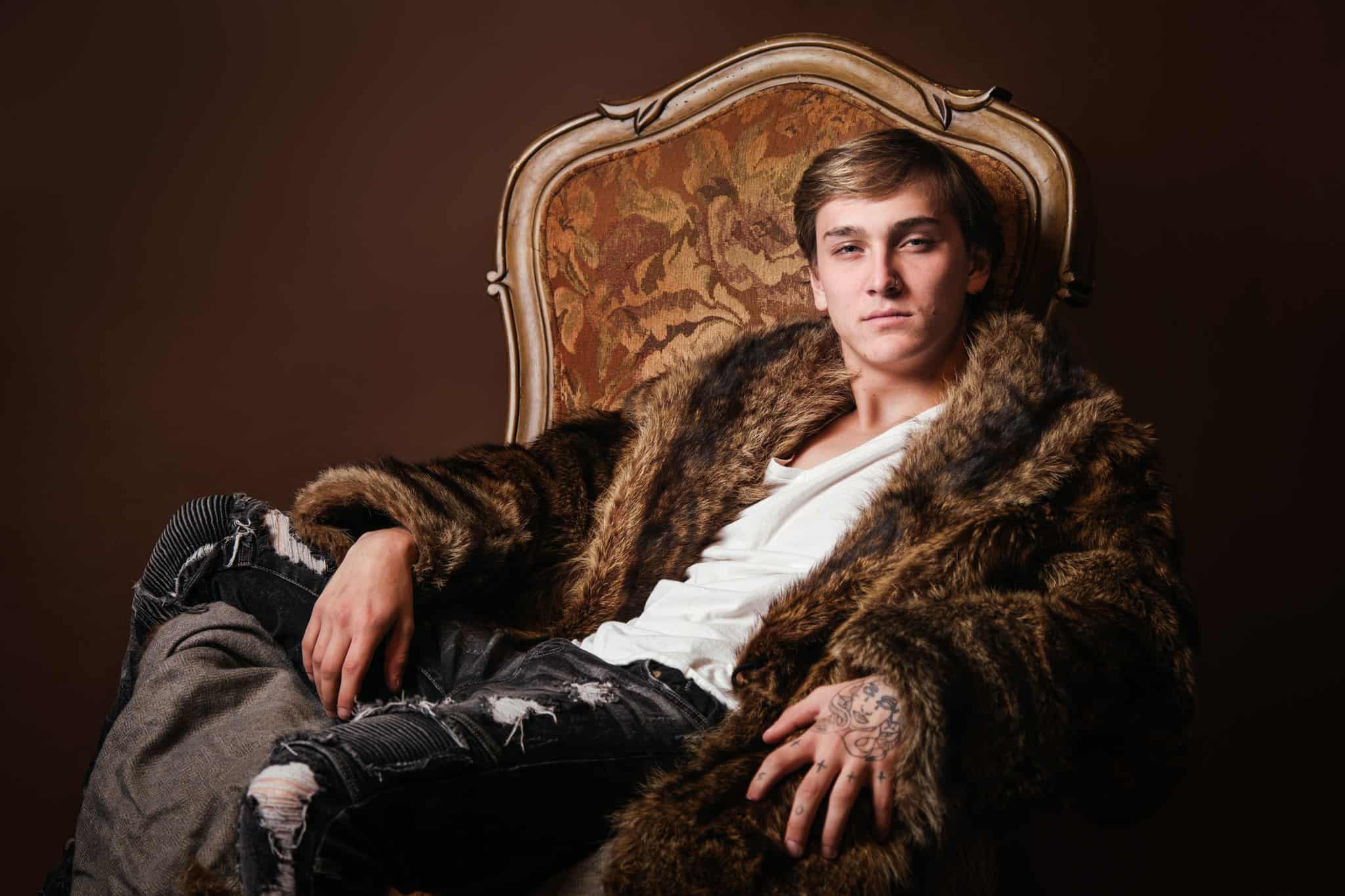 senior pictures for guys - portrait of young man in a fur coat sitting in a throne